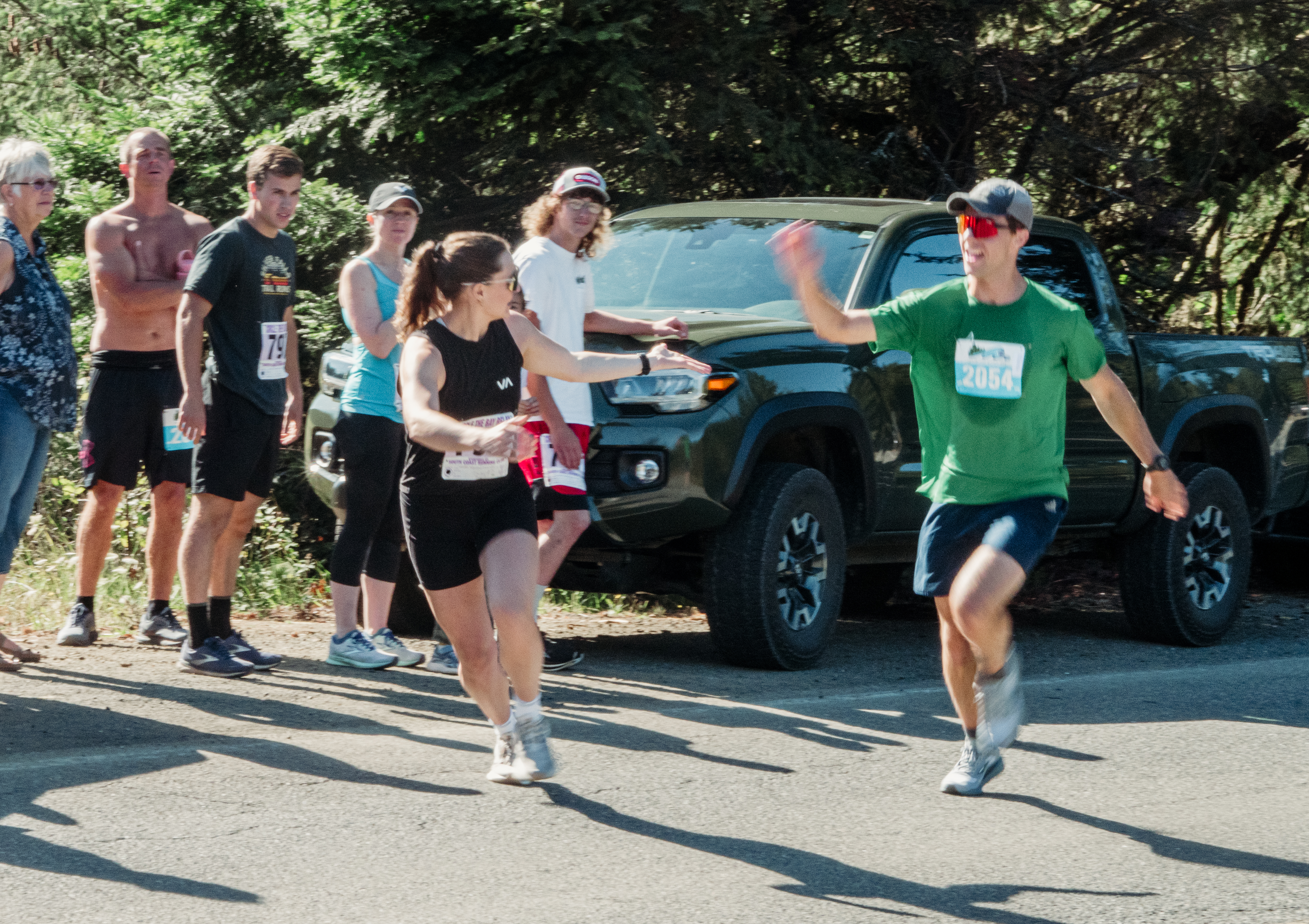 Two runners tag each other mid-stride during a race on a sunny day, while spectators stand along the roadside near a parked pickup truck, cheering them on.