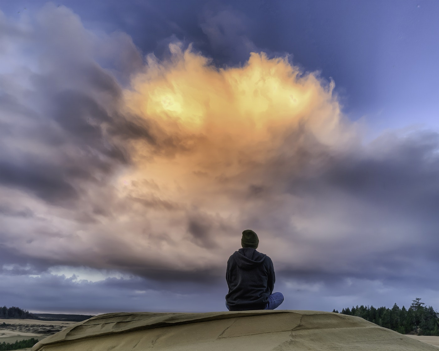 A person sits alone on top of a sand dune at the Oregon Dunes, gazing up at a dramatic sky with a large, glowing golden cloud formation surrounded by sweeping storm clouds at sunset.