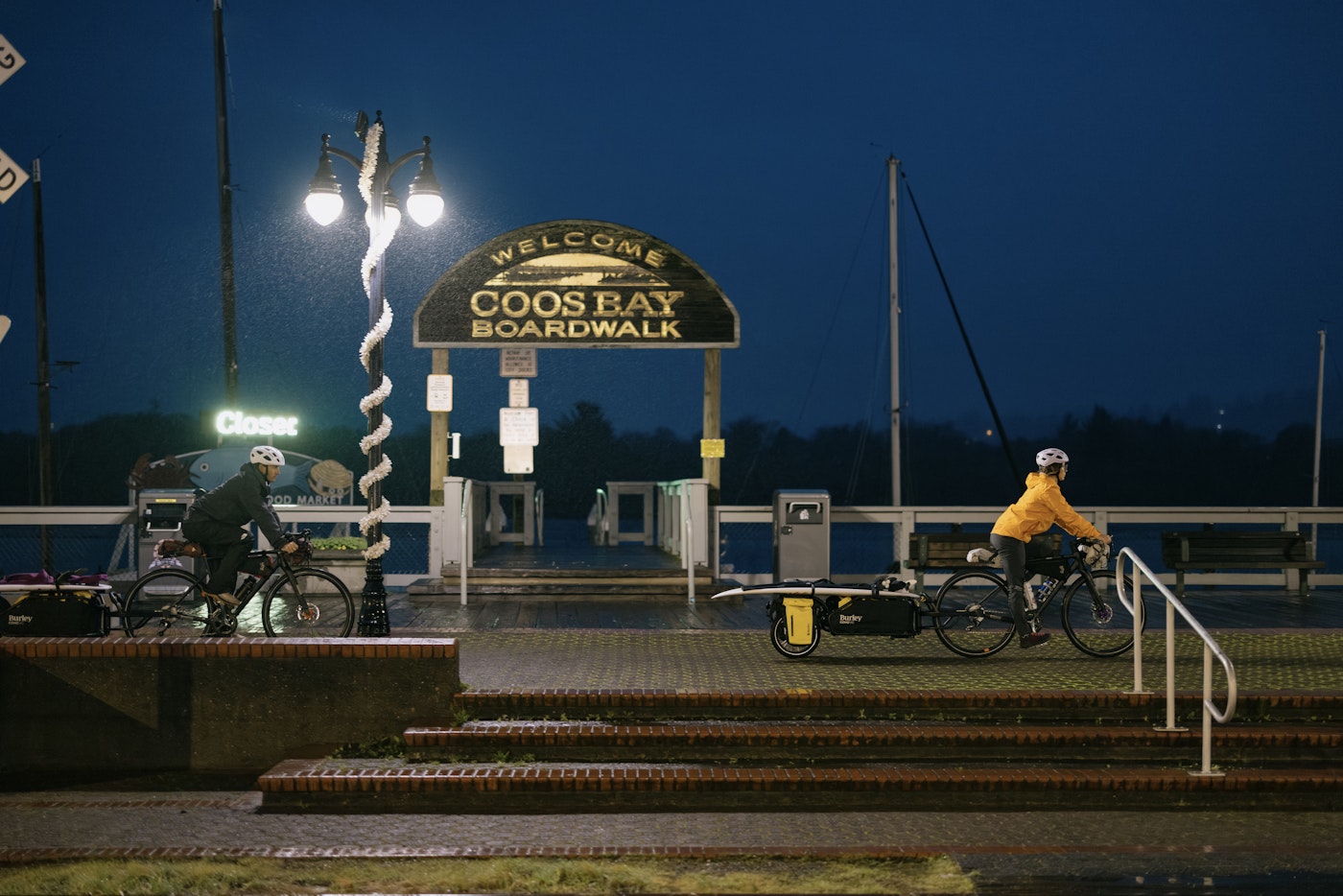 Two cyclists towing loaded cargo trailers ride past the lit Coos Bay Boardwalk sign on a rainy evening.