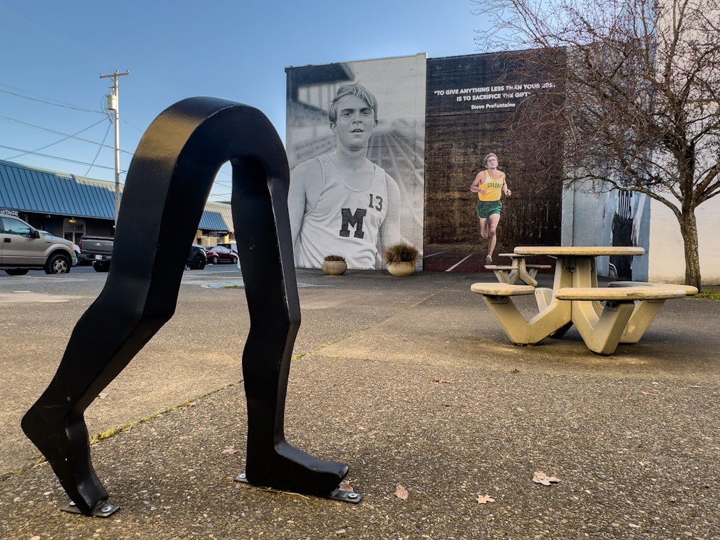 Black metal “Running Man” bike rack shaped like a runner’s legs installed in a small plaza in downtown Coos Bay, with the Steve Prefontaine memorial mural visible on the building wall in the background. Black metal “Running Man” bike rack shaped like a runner’s legs installed in a small plaza in downtown Coos Bay, with the Steve Prefontaine memorial mural visible on the building wall in the background.