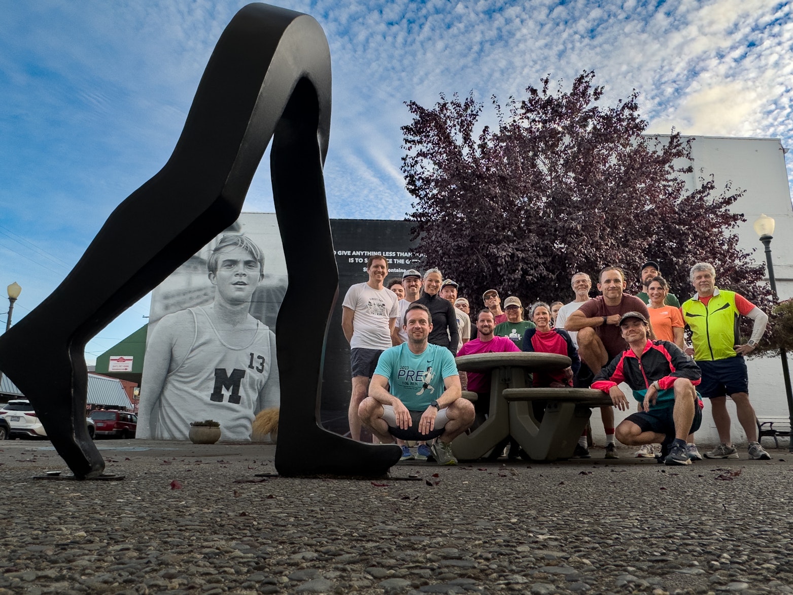 A group of runners poses for a photo beside a large abstract running sculpture, with a mural of Steve Prefontaine on a nearby building in the background.