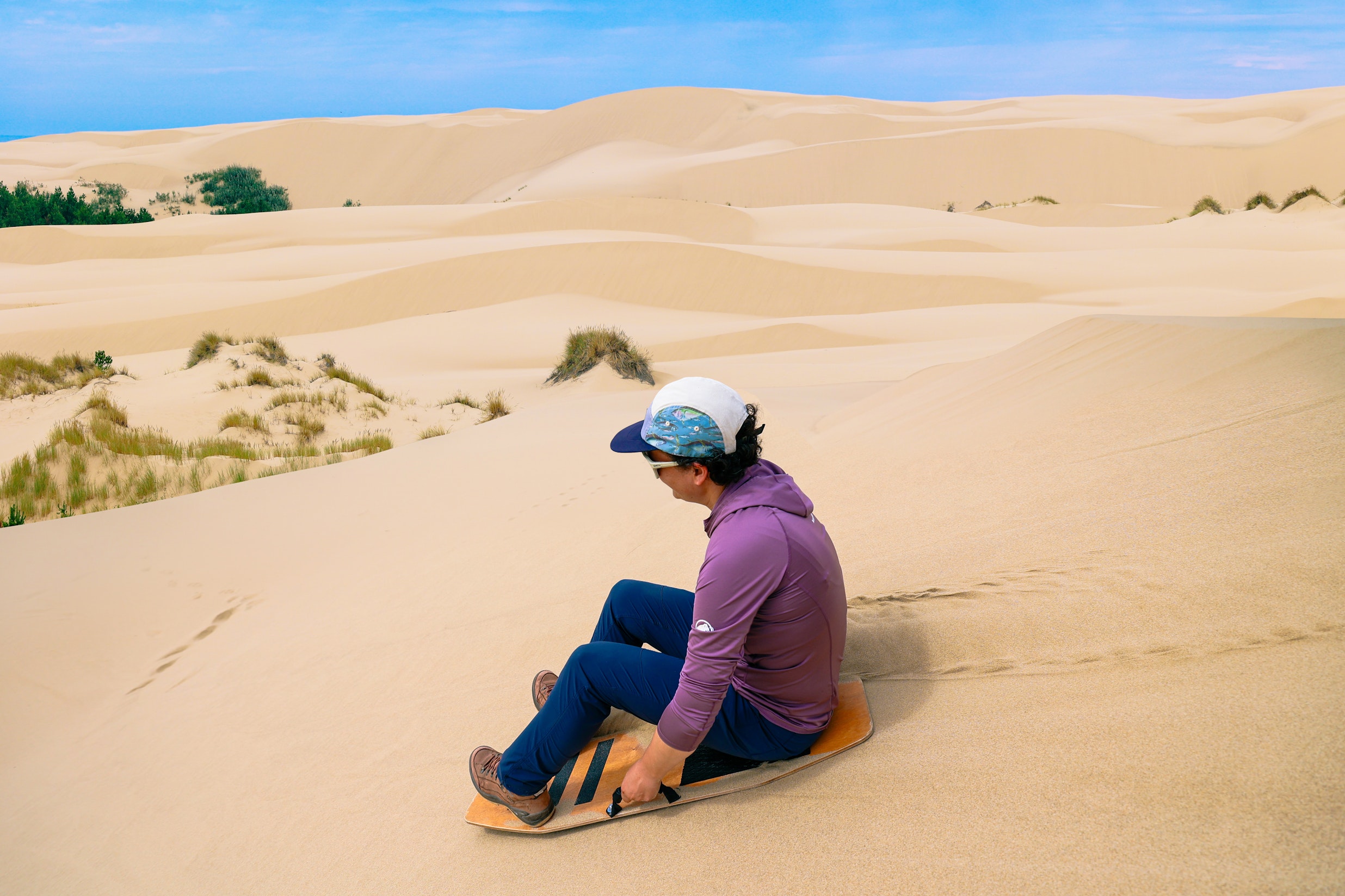 Person sandboarding down a large dune, seated on the board mid-descent with rolling dunes stretching into the distance.