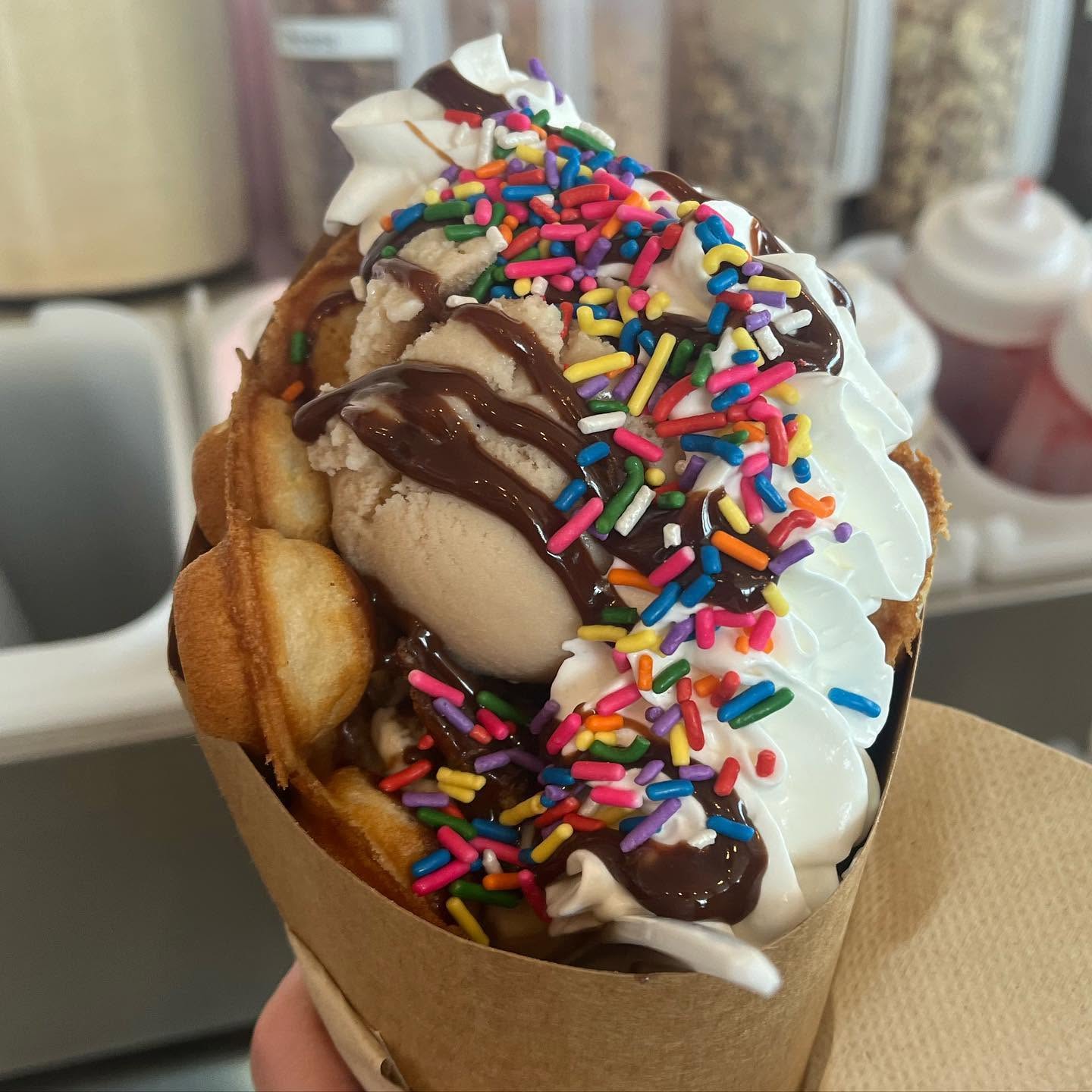 Image courtesy of Scoops Handmade Ice Cream A photo of Scoops' bubble waffle cone filled with ice cream, whipped cream, chocolate drizzle, and rainbow sprinkles, held in a paper wrap.