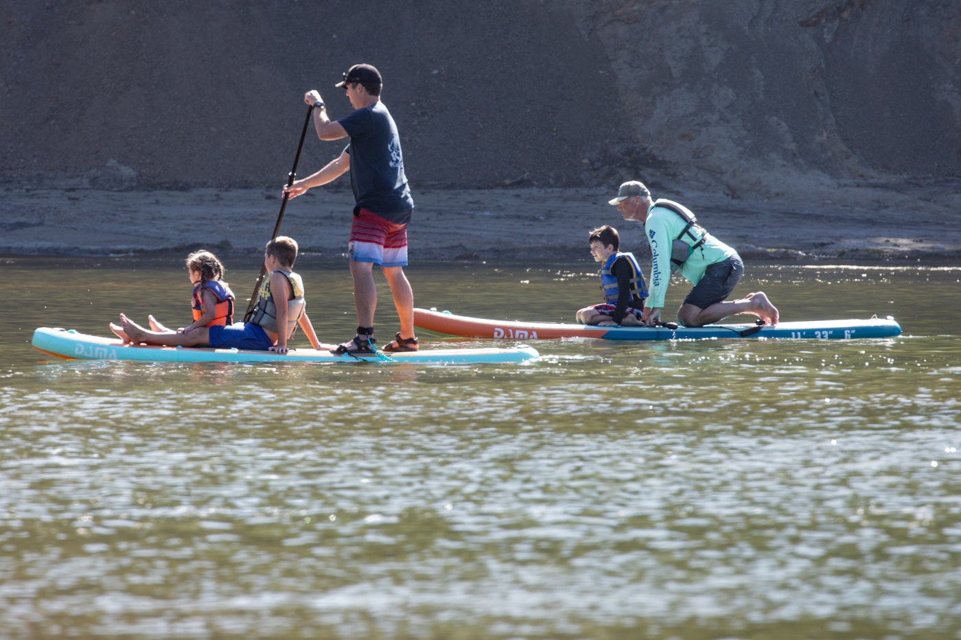 Two adults paddleboarding on calm water with three children wearing life jackets seated on the boards, with a rocky bluff in the background.