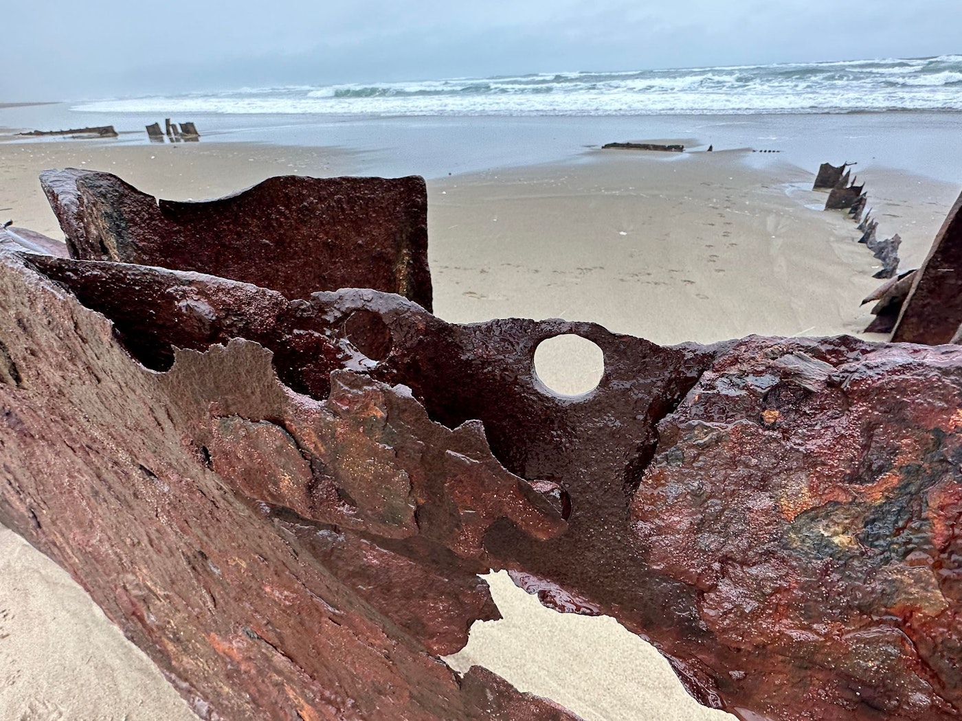 Close-up of large rusted, weathered metal shipwreck wreckage from the 1929 wreck of the Sujameco, jutting from Horsfall Beach, with more scattered wreckage fragments and ocean waves in the background.