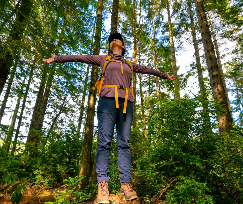 A hiker wearing a backpack stands on a forest trail with arms outstretched, surrounded by tall evergreen trees and lush greenery.