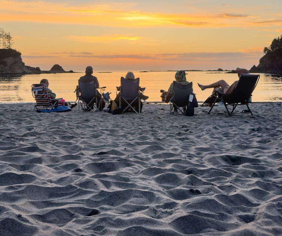 A group of people sit in beach chairs on the sand, facing the ocean and watching a calm sunset over the water.