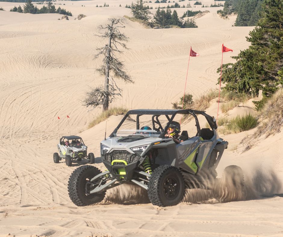 Two off-road vehicles drive across rolling sand dunes, kicking up sand as riders navigate the open landscape marked with safety flags.