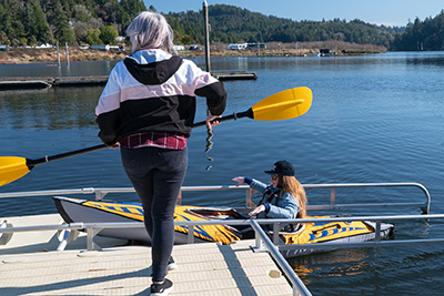 Woman on dock holding kayak paddle with woman in kayak on the water