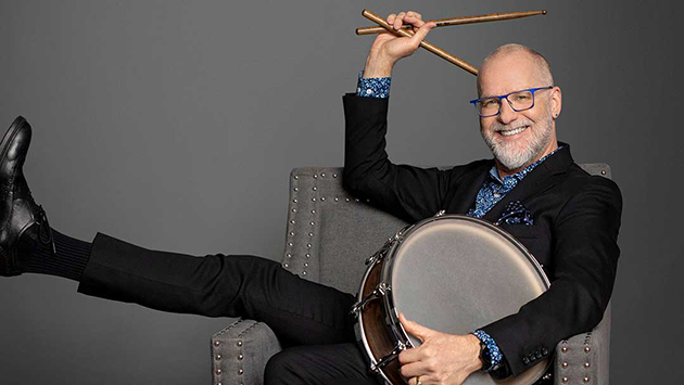 Percussionist Terry Longshore sitting in a grey chair holding a drum and drum sticks.