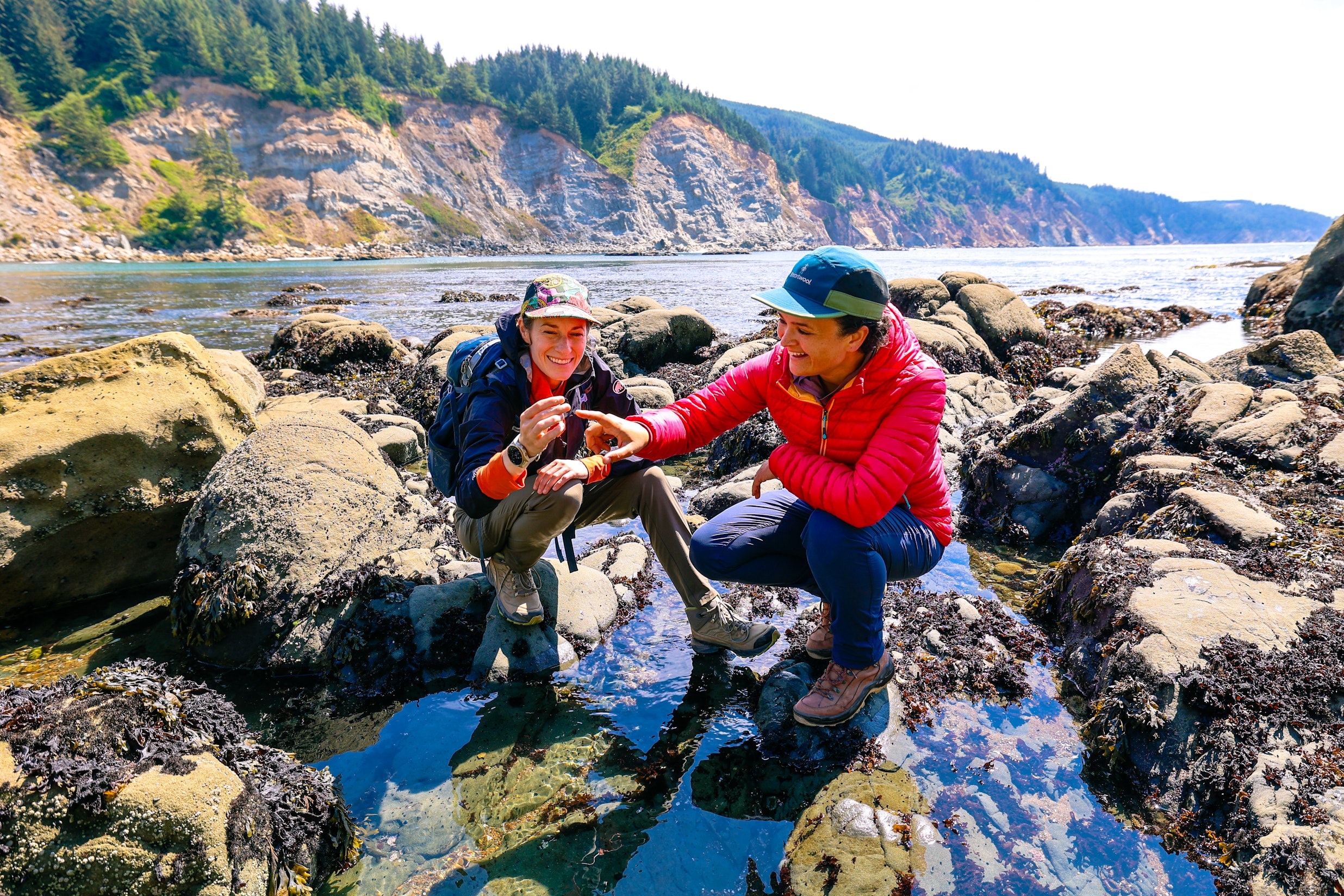 Photo @ Gwyn and Ami Two women in hiking gear crouch among tidepools on a rocky coastline, smiling as they examine a small find, with rugged cliffs and forested hills in the background.