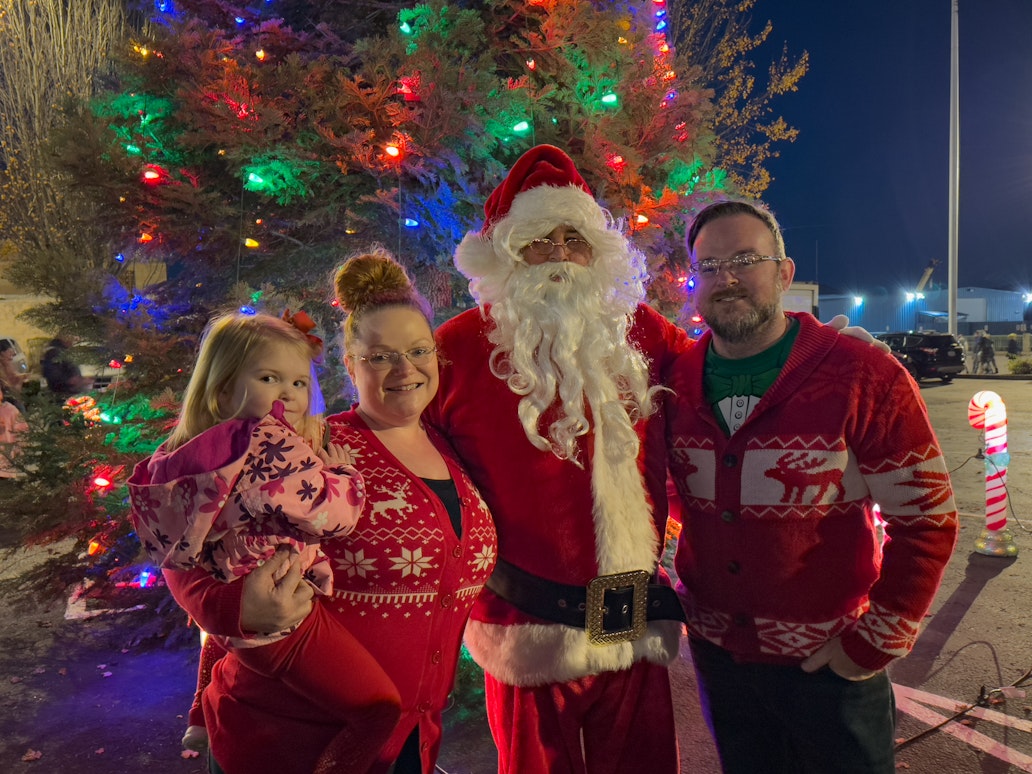 A family poses with Santa in front of a brightly lit Christmas tree during a tree lighting event in Coos Bay, where visitors enjoy photos with Santa, holiday treats, caroling, and festive community activities.